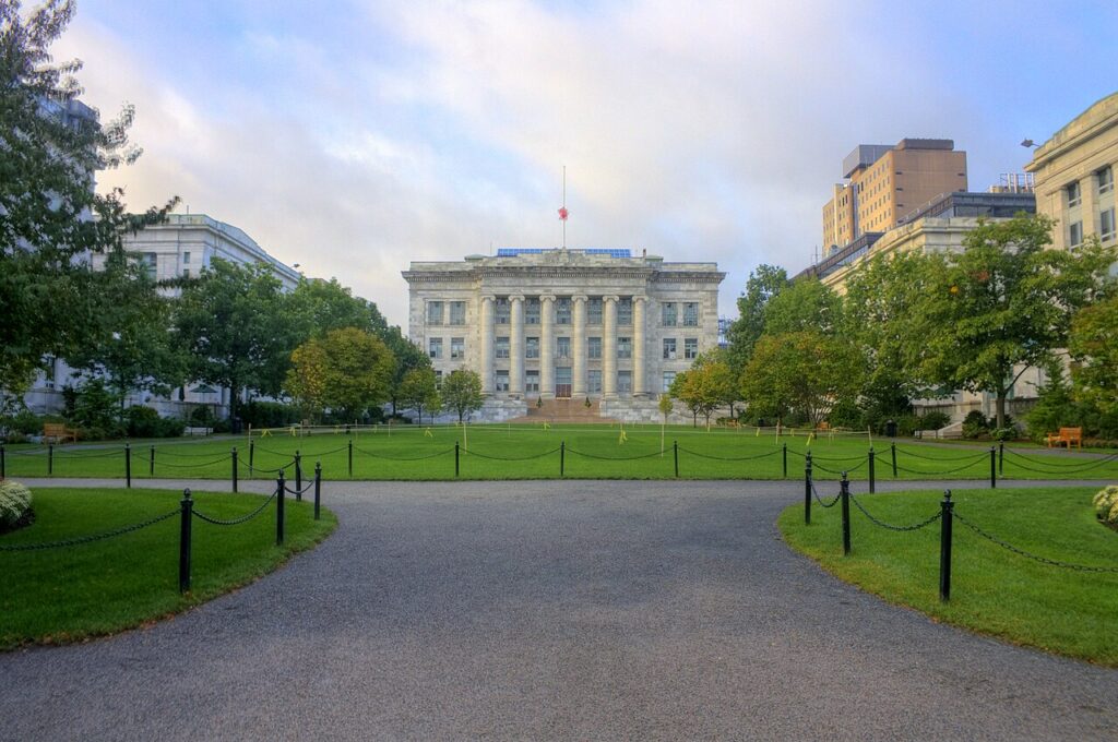 Harvard University's Widener Library viewed across Harvard Yard, showing the iconic neoclassical building with its prominent columned facade and wide front steps. A tree-lined pathway leads across the manicured green lawn toward the library, with the walkway bordered by low black chain fencing. Mature trees frame the scene on both sides, and other Harvard buildings are visible in the background under a partly cloudy sky.